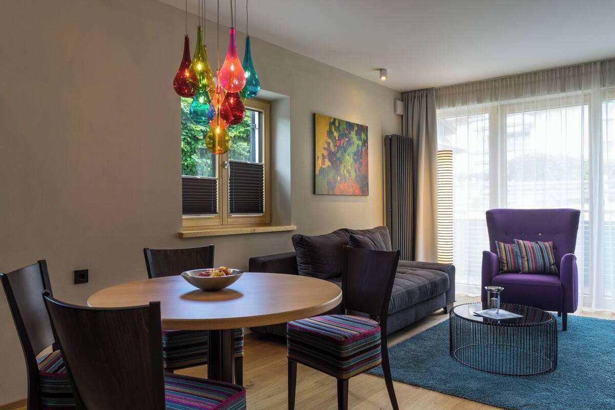 Una sala de estar moderna con una mesa de comedor redonda, luces colgantes coloridas, un sillón morado y un ventanal con persianas.
