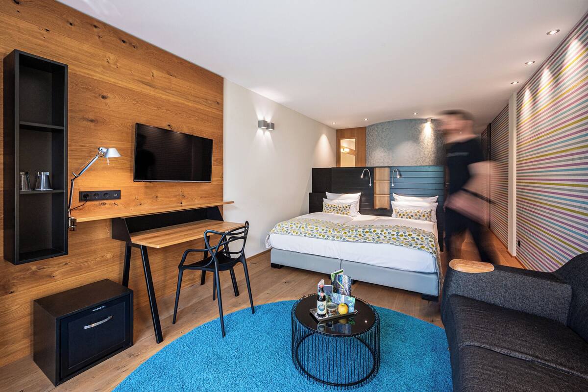 Una habitación de hotel moderna con una pared de madera, un televisor de pantalla plana, un escritorio, una cama con edredón estampado, una mesita de café y un sofá.