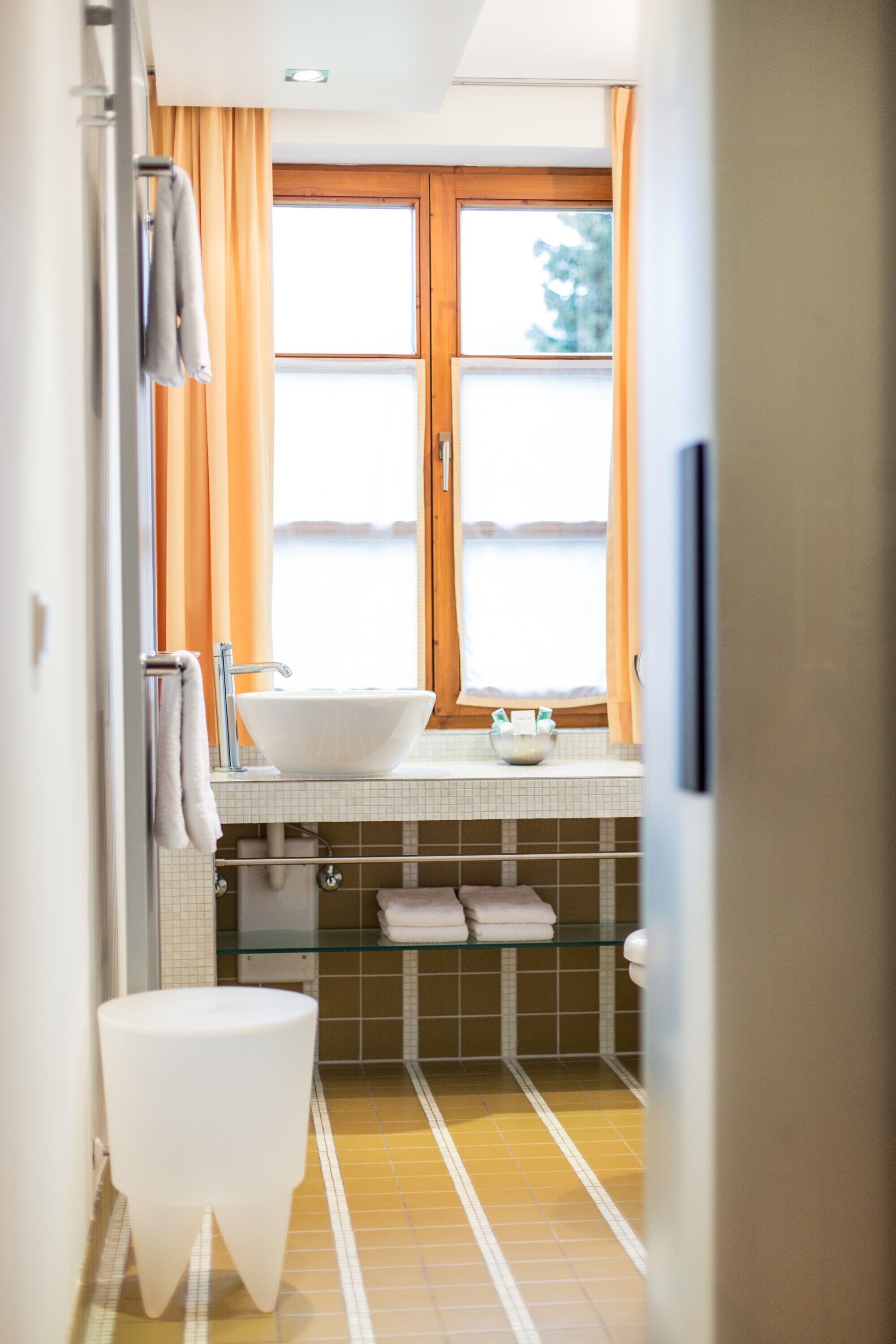 A bathroom with a white sink, a glass shower enclosure, and a window with orange curtains.