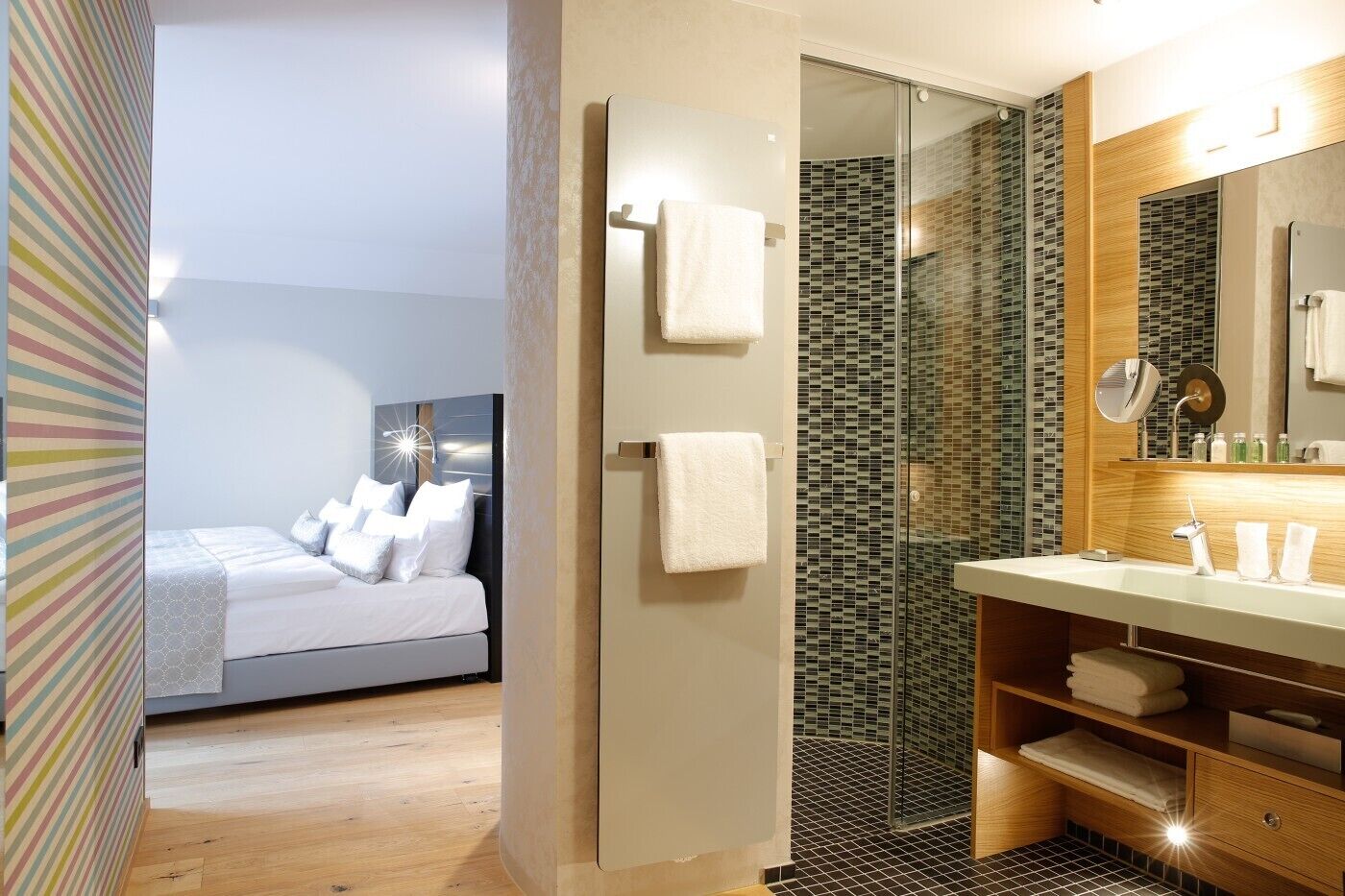 A modern hotel room with a bed, a shower area with glass doors, and a vanity with a mirror and toiletries.