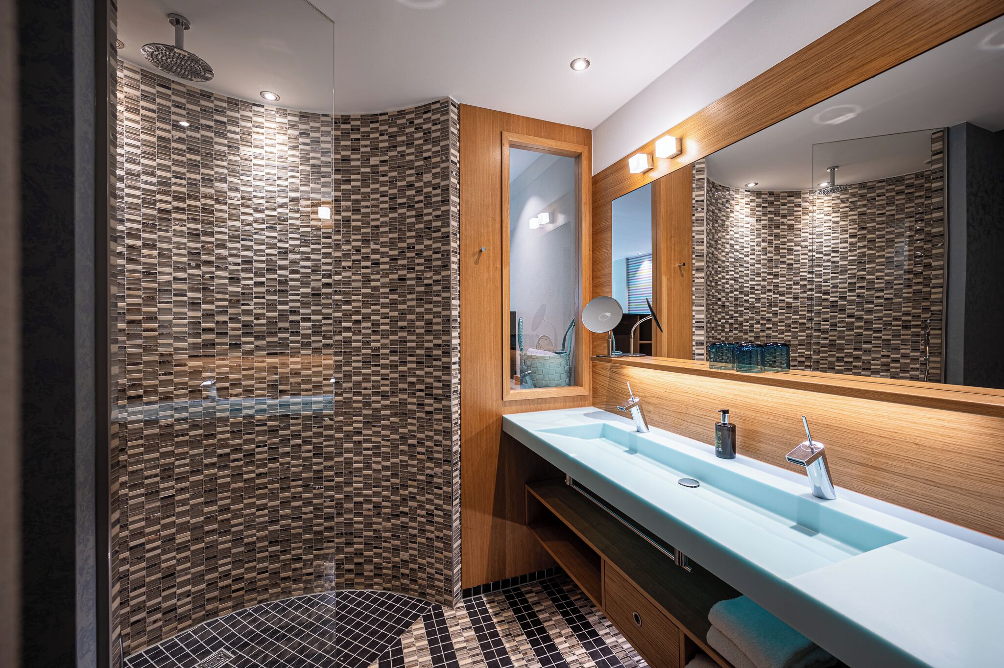 Une salle de bain moderne avec une douche à l’italienne en verre, un meuble-lavabo en bois doté d’un grand miroir et un lavabo bleu clair.