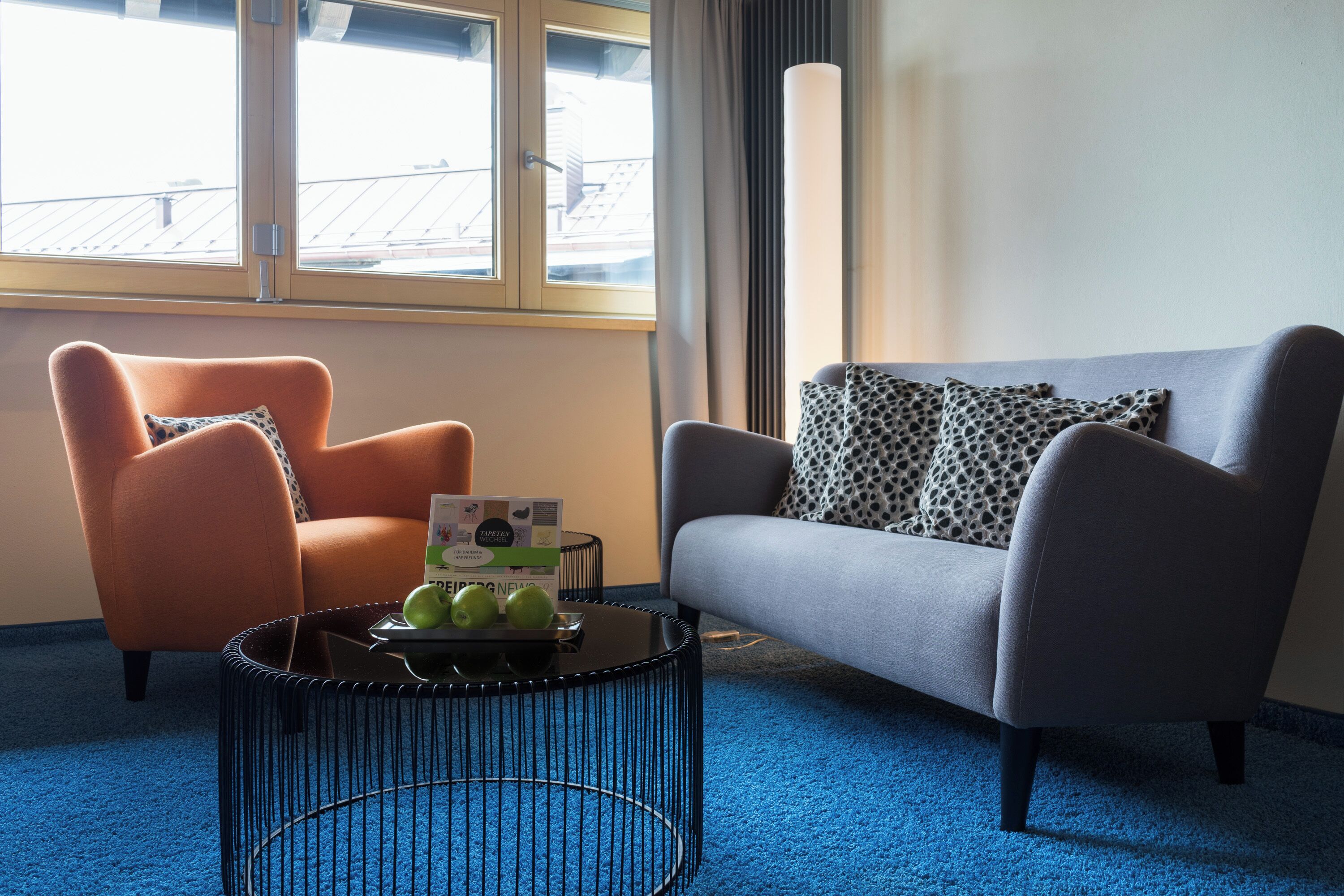 A room with a blue carpet, a grey sofa, an orange armchair, and a black coffee table with a magazine and apples.