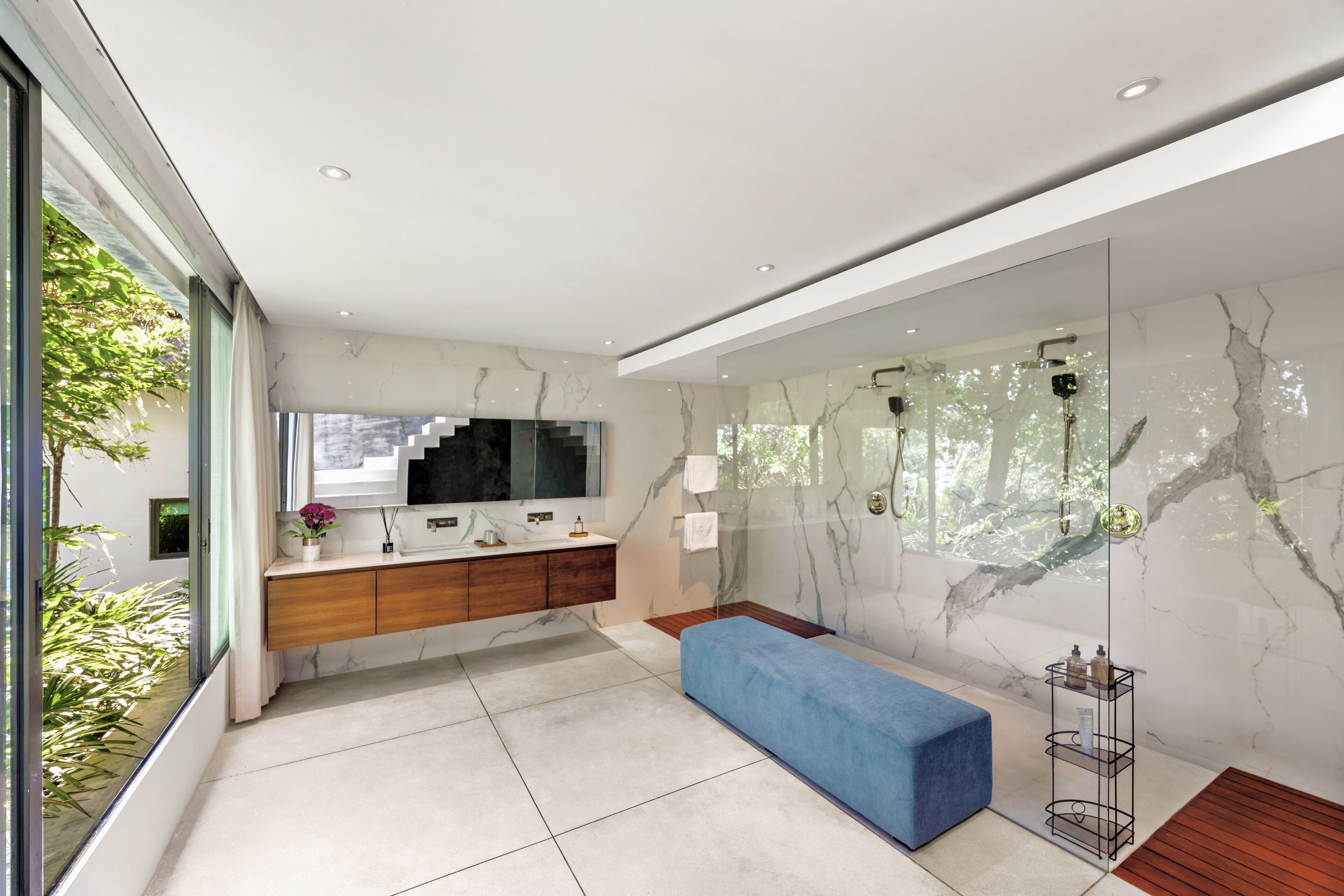 A modern bathroom with a marble wall, a large mirror, a wooden vanity, a blue bench, and a glass shower enclosure.
