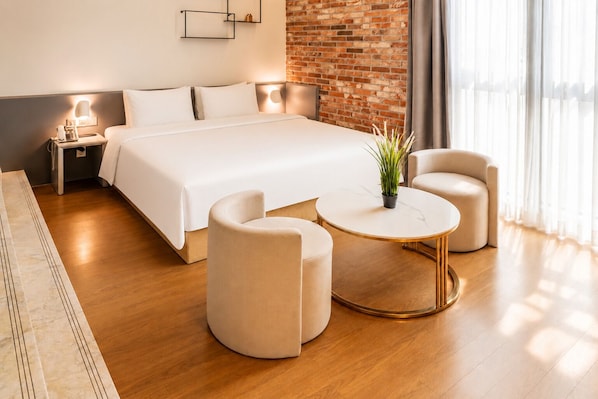 Una habitación de hotel moderna con una cama grande, dos sillones, una pequeña mesa redonda, una lámpara de noche y una pared de ladrillo.