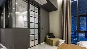 Quarto de hotel moderno com porta de vidro deslizante, um pequeno sofá, uma mesa de madeira e vista para a cidade à noite.