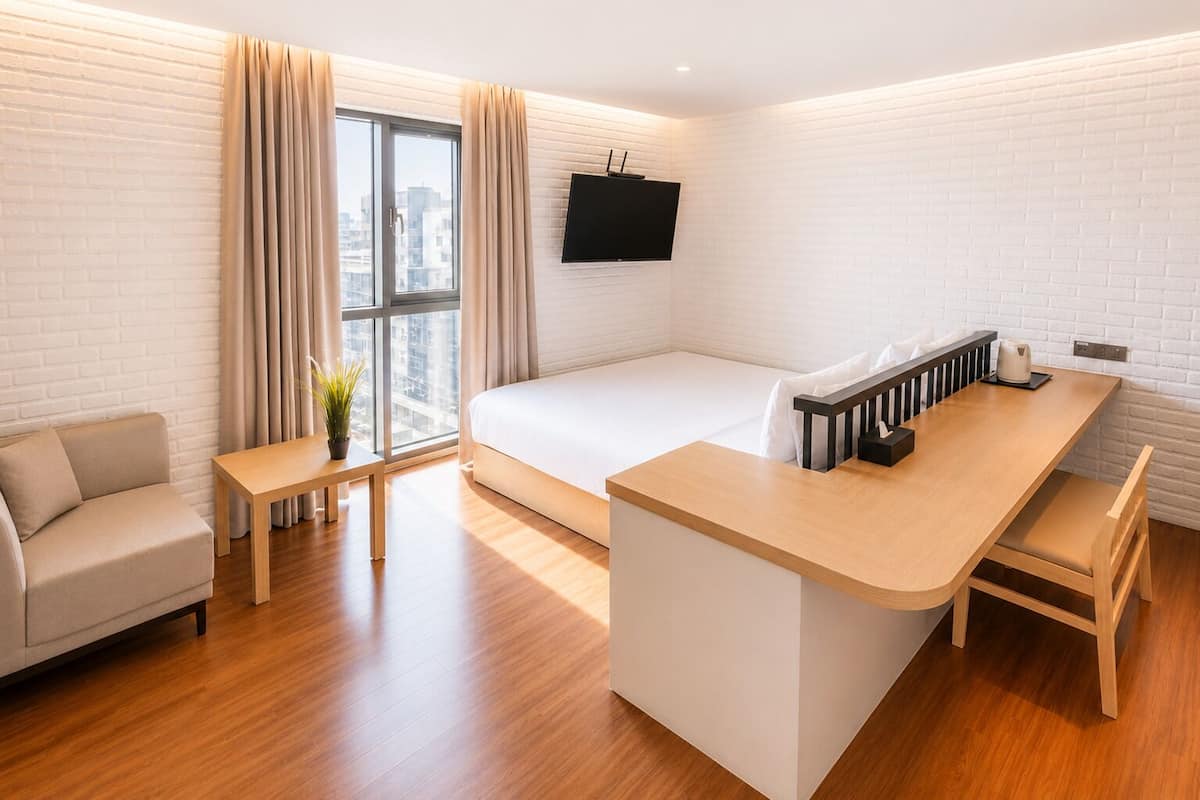 Quarto de hotel moderno com cama, sofá, mesa pequena, escrivaninha e televisão fixada na parede.