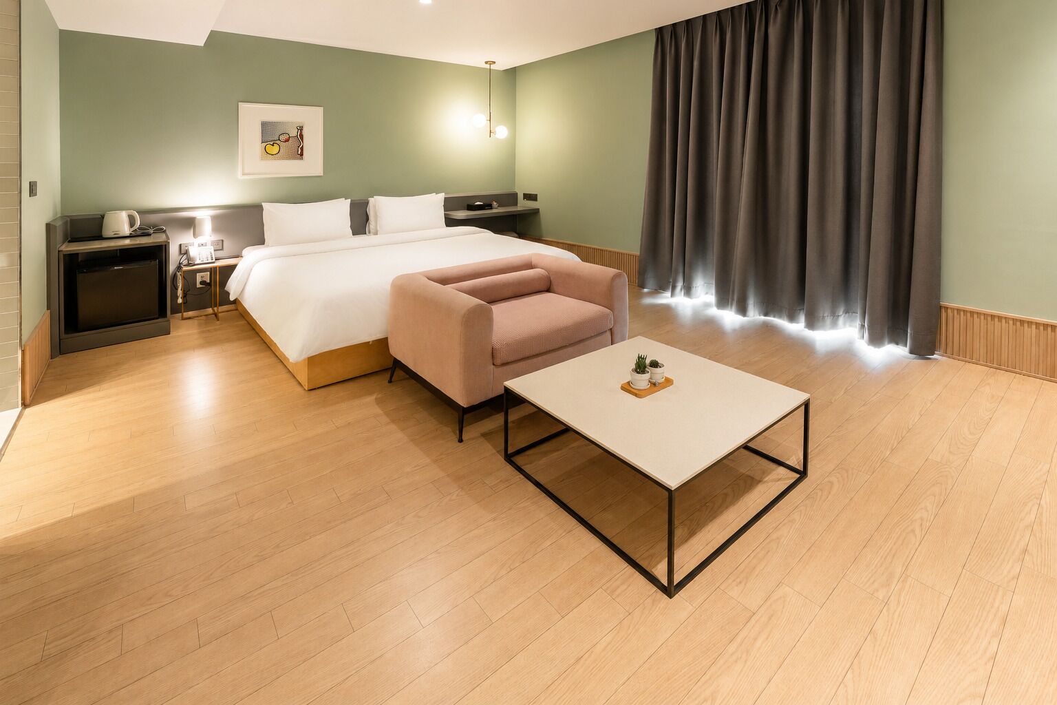 Habitación de hotel con cama, sofá, mesa de centro, mesita de noche, lámpara y un cuadro en la pared.