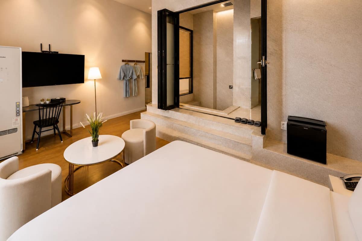 Quarto de hotel com uma cama, uma escrivaninha iluminada por um abajur, uma cadeira, uma pequena mesa com uma planta e uma televisão fixada na parede.