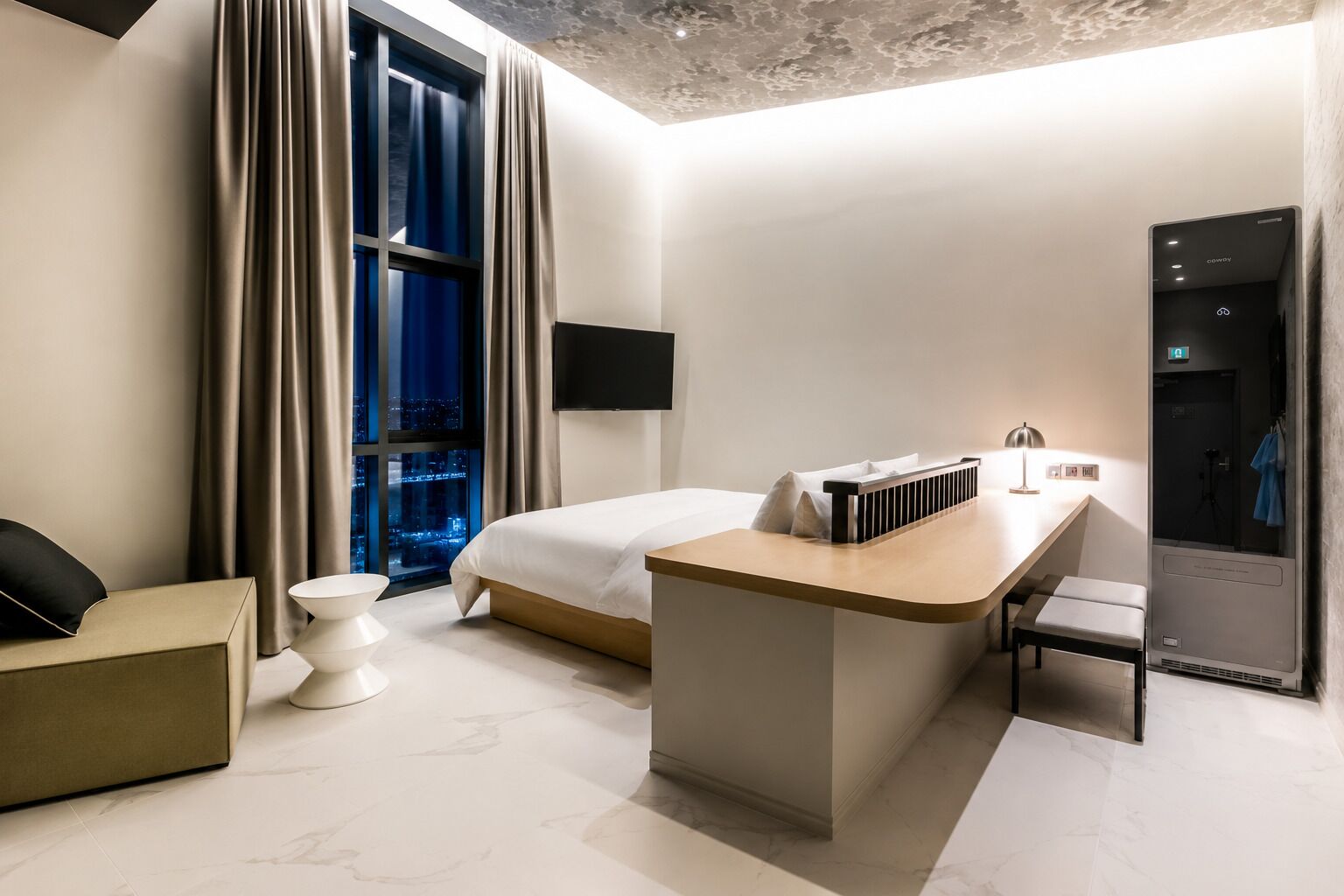 A modern hotel room with a bed, a desk, a chair, a small table, and a view of the city at night.