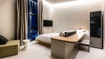 A modern hotel room with a bed, a desk, a chair, a small table, and a view of the city at night.