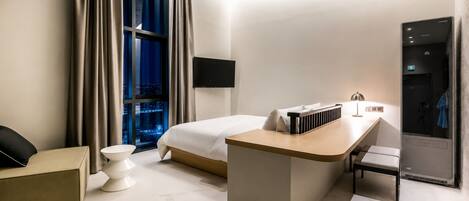A modern hotel room with a bed, a desk, a chair, a small table, and a view of the city at night.