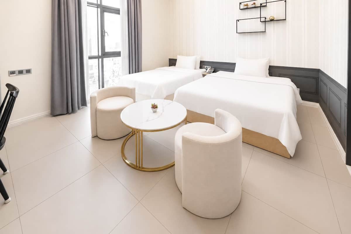 Quarto de hotel com cama, duas poltronas, uma mesa redonda pequena e vista da cidade por grandes janelas.