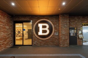 Entrada de un hotel con un gran letrero iluminado con la letra 'B', fachada de ladrillo y una recepción visible a través de las puertas de vidrio.
