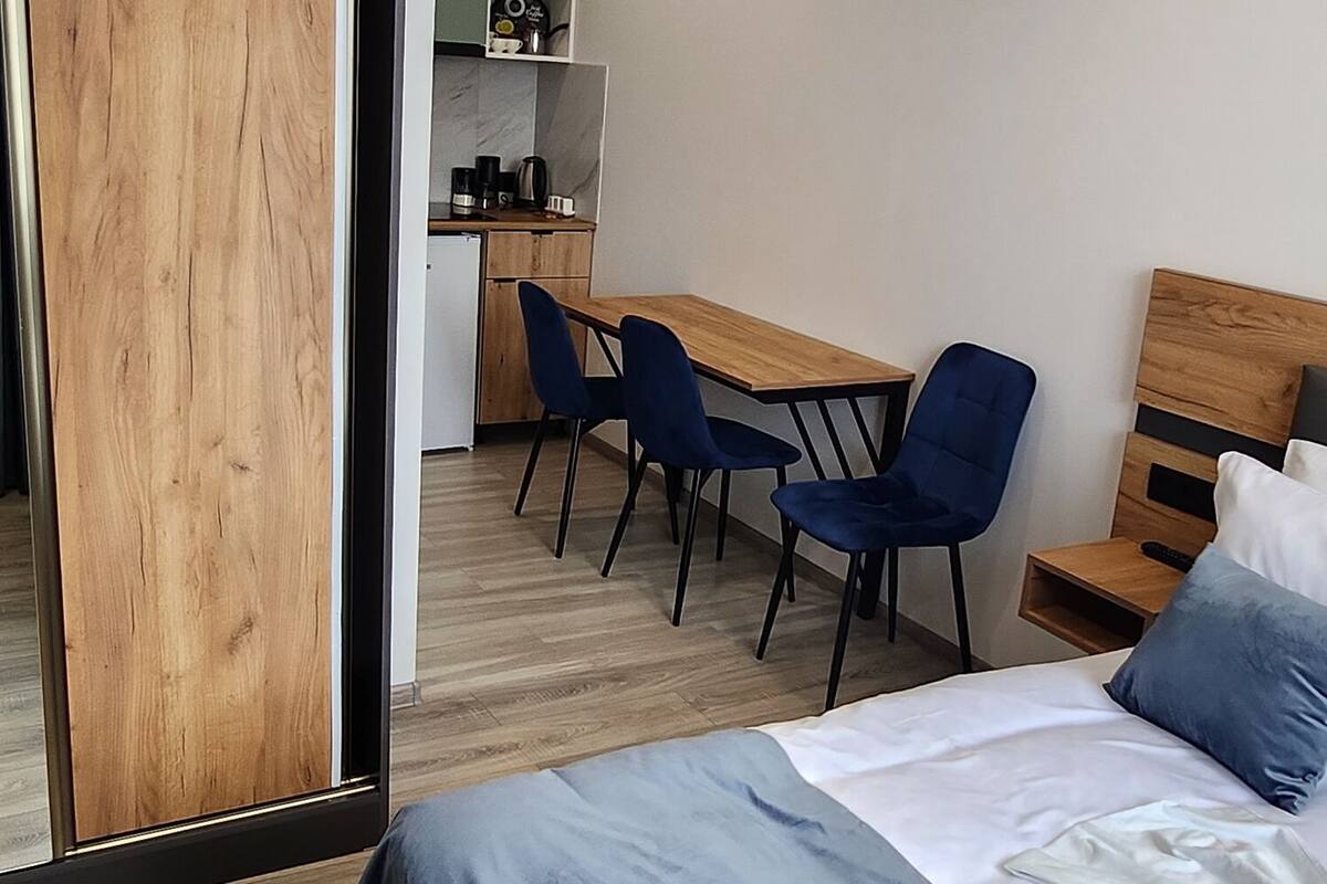 Ein modernes Schlafzimmer mit einem Holzbett, einem Schreibtisch, Stühlen und einer kleinen Küchenzeile im Hintergrund.