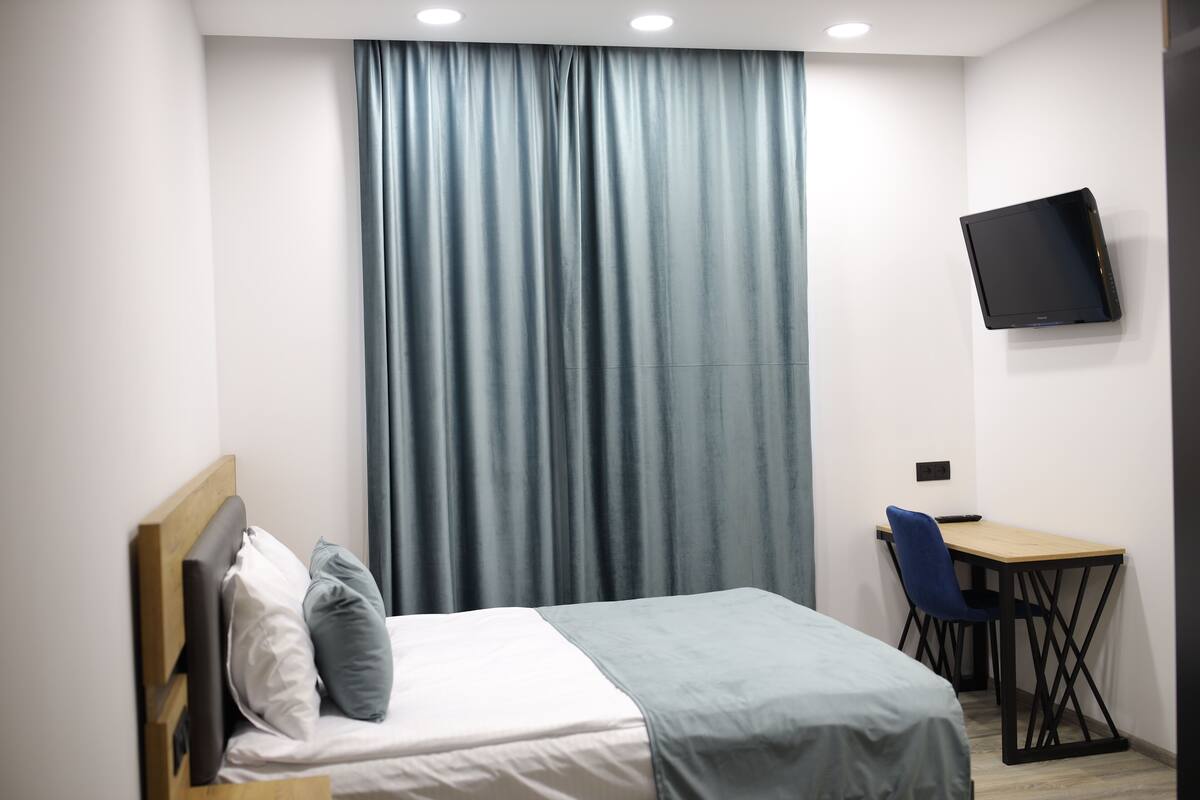 Ein Schlafzimmer mit Bett, Schreibtisch, Stuhl, Fernseher und einem Fenster mit Vorhängen.