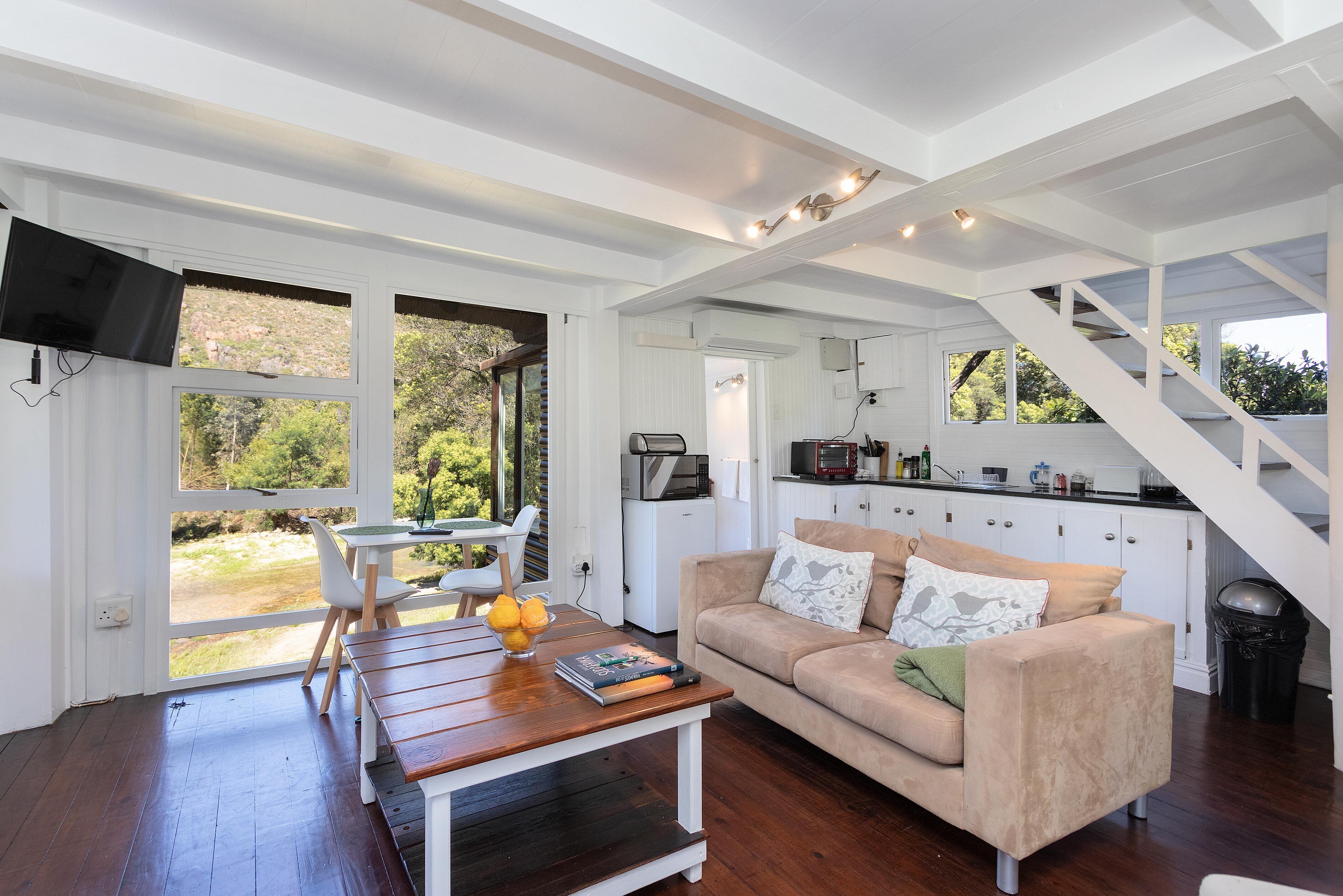 A spacious living room with a wooden coffee table, a beige sofa, and a dining area with a table and chairs. A kitchen area is visible in the background with white cabinets and a staircase leading upstairs.