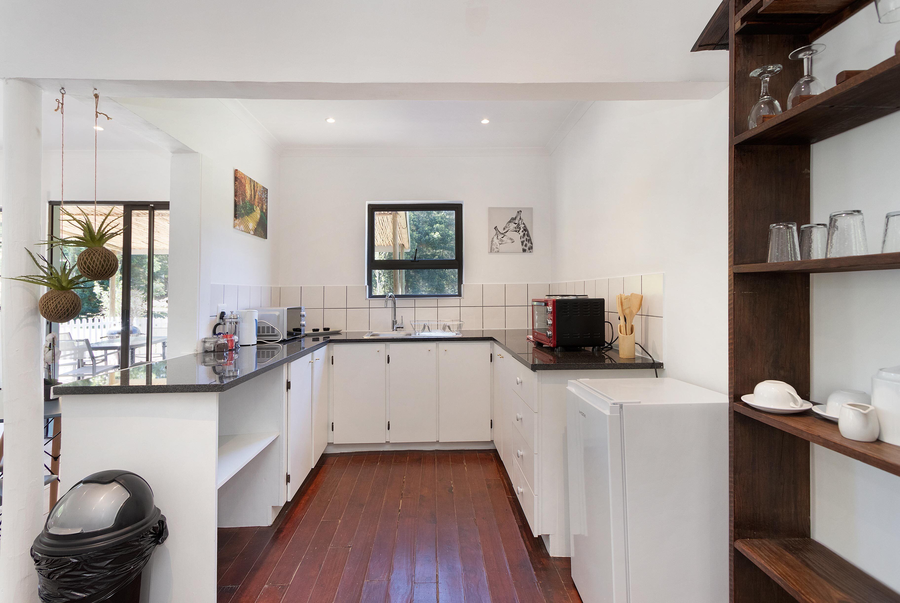 A modern kitchen with white cabinets, a black countertop, and a wooden floor. There is a window with a view of greenery outside, and a shelf with glassware.