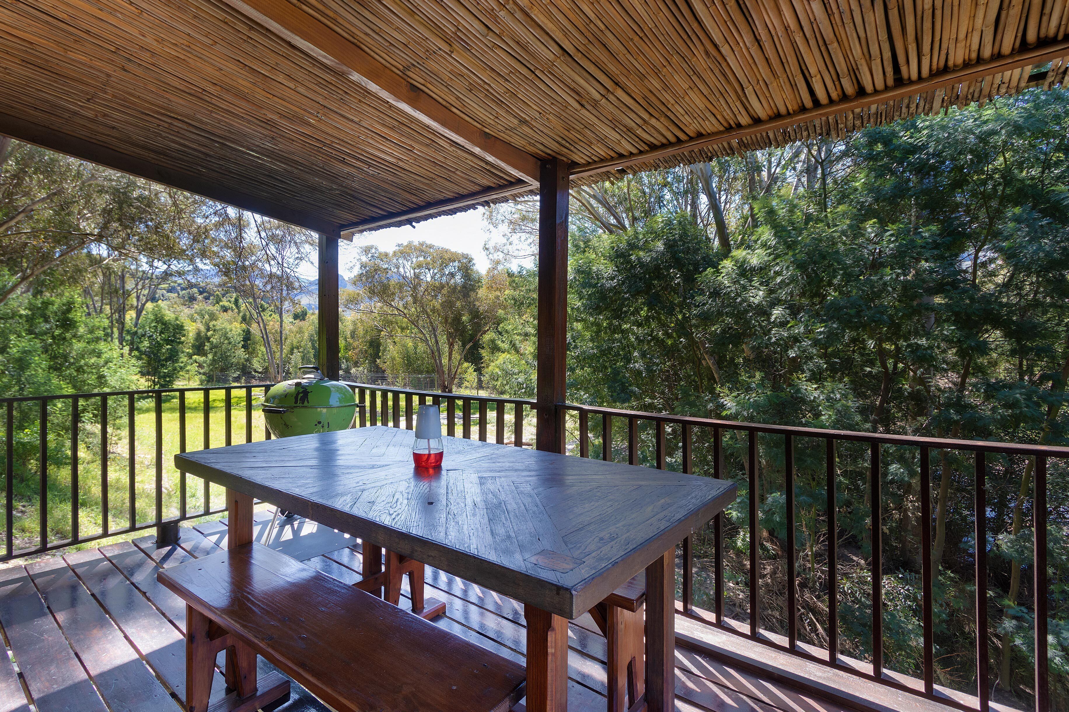 A covered outdoor area with a wooden table and benches, surrounded by a metal railing, overlooking a forested landscape.