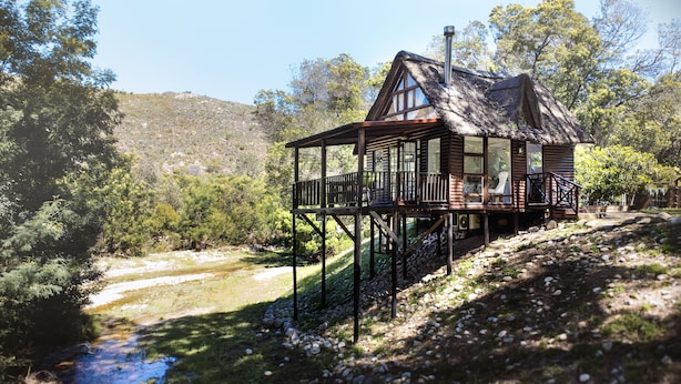 A wooden cabin with a balcony, situated by a river, surrounded by trees and hills. - La Ferme Guest Farm (Franschhoek)