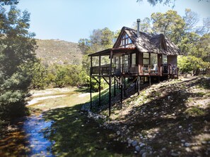 Une cabane en bois dotée d’un balcon, située au bord d’une rivière, entourée d’arbres et de collines.