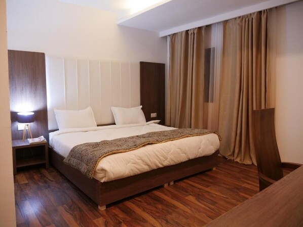 Habitación de hotel con una cama grande, suelo de madera, una mesita de noche con lámpara y vistas al exterior a través de cortinas.