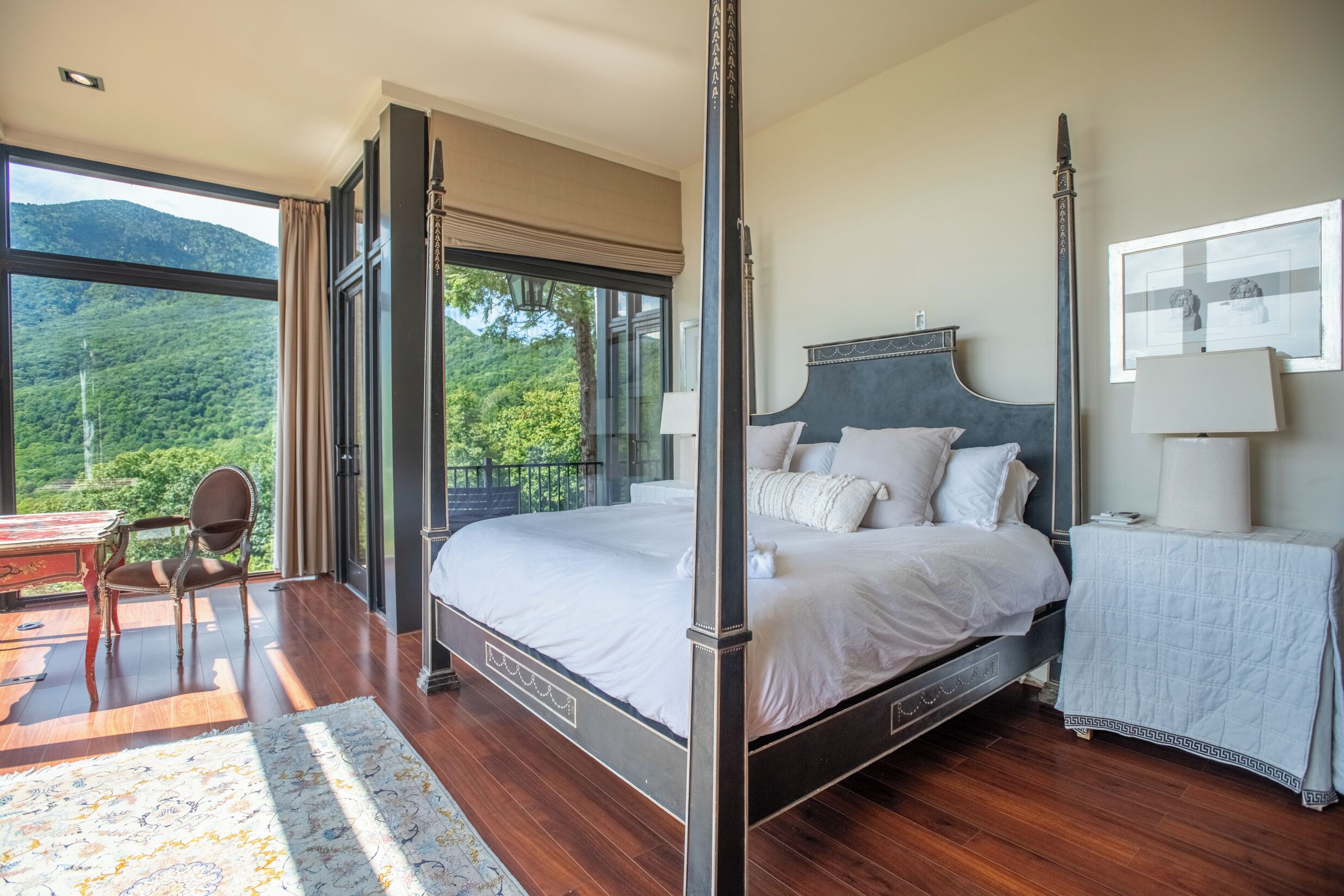 A bedroom with a four-poster bed, a large window with a view of a forest, a desk with a chair, and a nightstand with a lamp.
