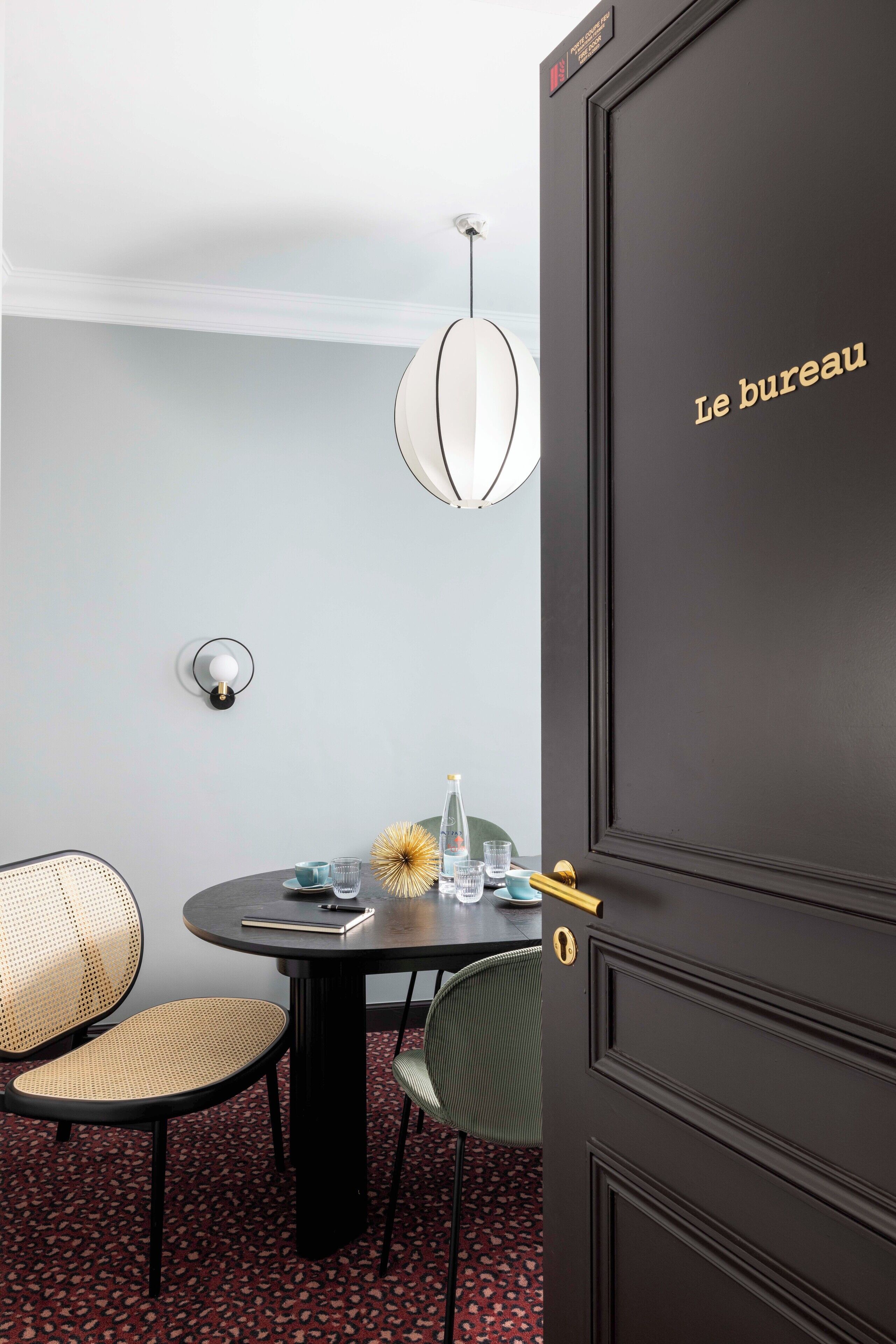 A room with a dark wooden door labeled "Le bureau," a round table with chairs, a hanging light fixture, and a wall-mounted light.