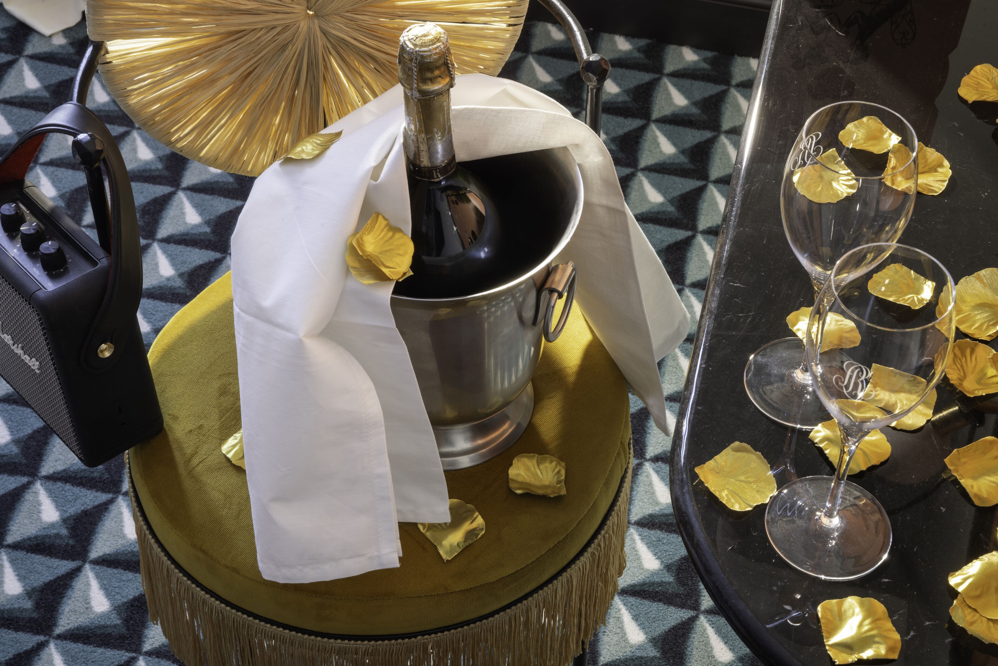 Une bouteille de champagne dans un seau métallique, entourée d’éléments décoratifs dorés et blancs sur une nappe à motifs.