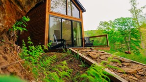 A modern cabin with a large glass window, wooden exterior, and a deck. - Unique Cliff Hot Tub with Views | Mural | Sleeps 4 (Rogers)