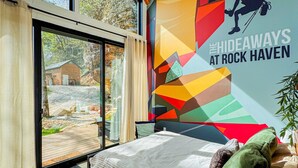 A modern bedroom with a large window, a bed, and a colorful wall mural. - Unique Cliff Hot Tub with Views | Mural | Sleeps 4 (Rogers)