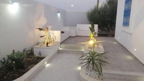 Un patio con paredes blancas, suelo de baldosas y varias plantas en macetas. Hay escalones que conducen a un nivel superior, y una pequeña mesa con una lámpara.