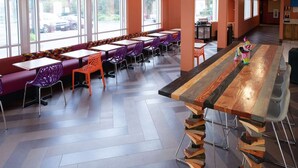 Un espace repas moderne avec de longues tables et des chaises colorées, de grandes fenêtres et une table centrale en bois.