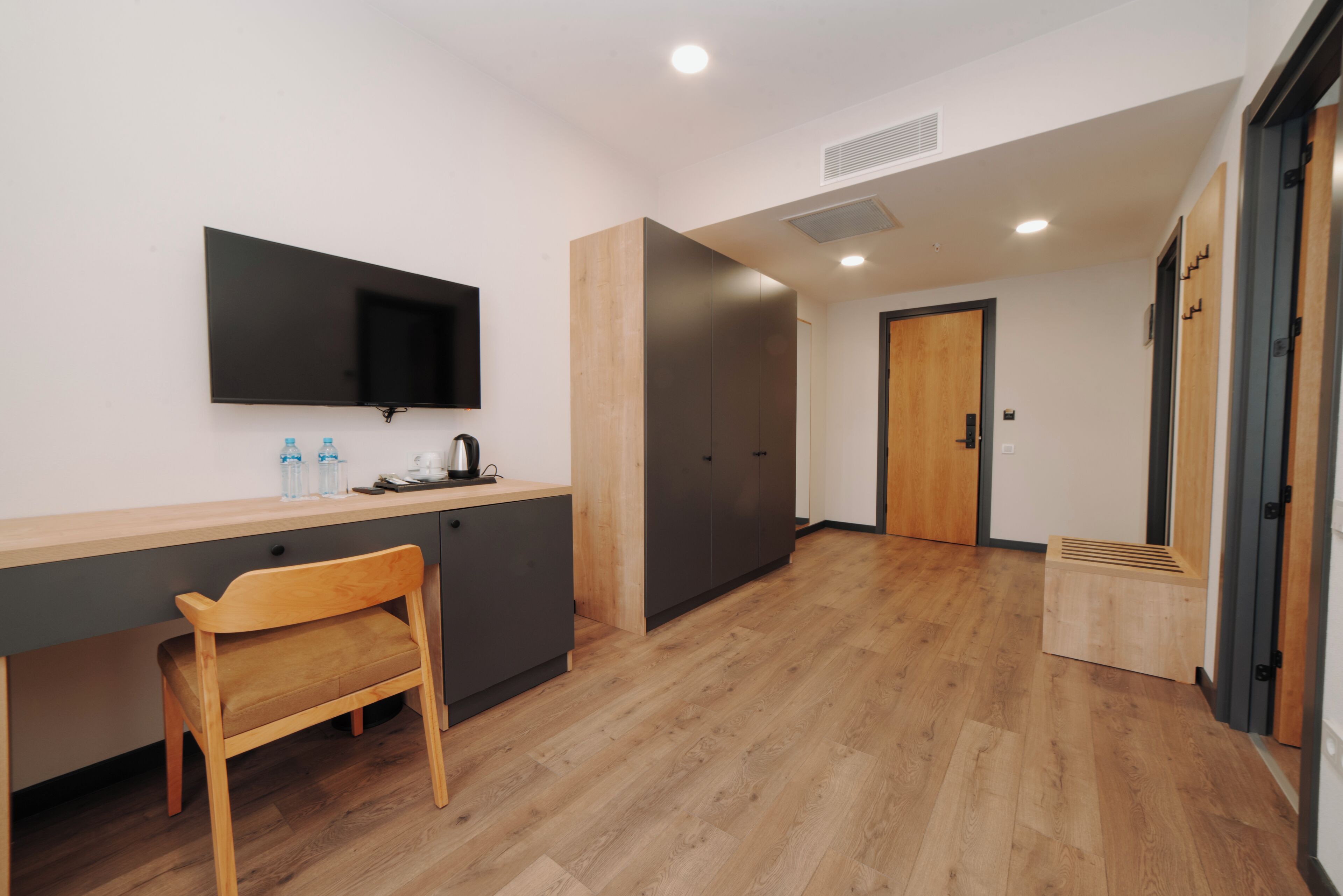 A modern, well-lit room with a wooden desk, a chair, a flat-screen TV mounted on the wall, and a door leading to another room.