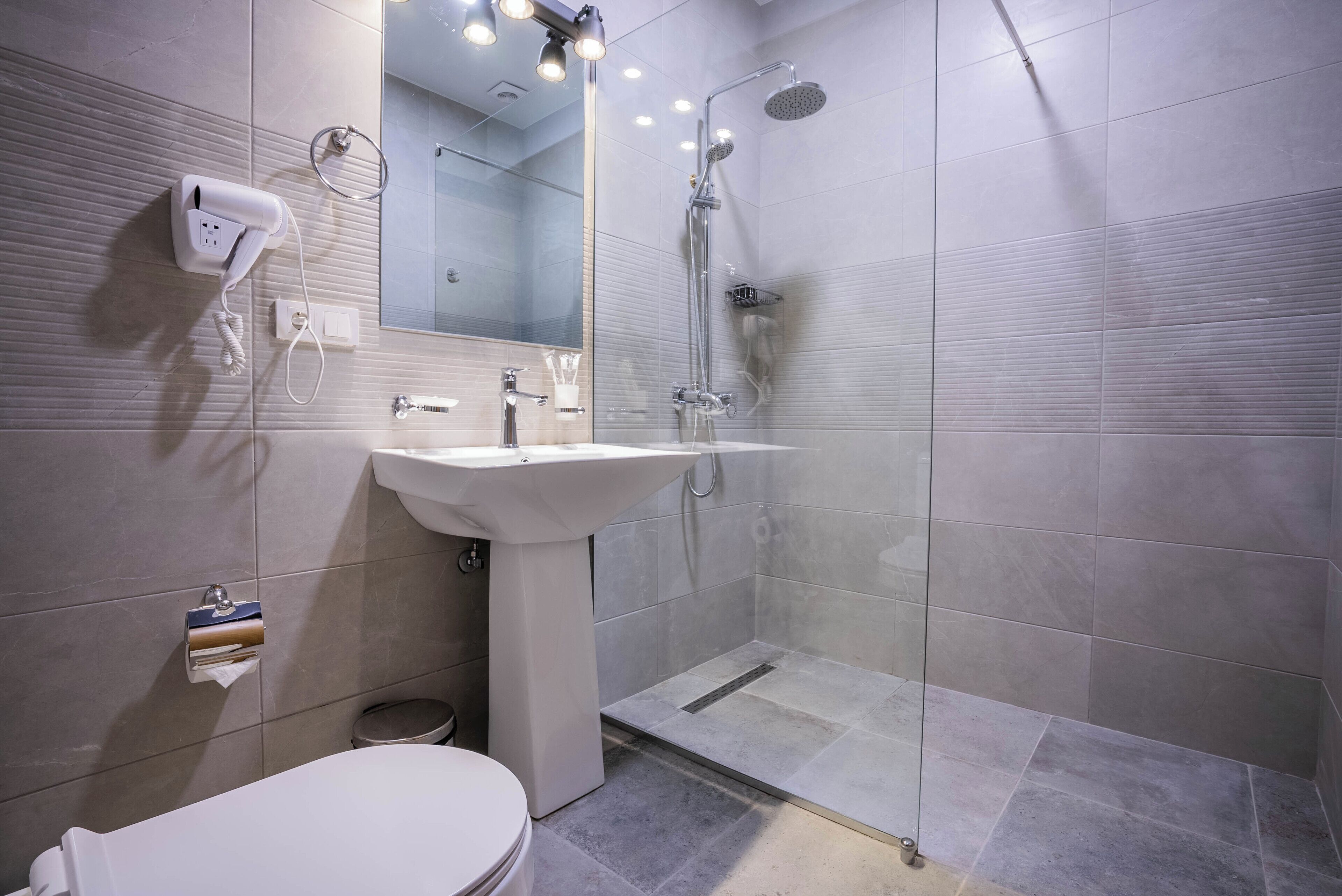 Un baño moderno con un lavamanos blanco, un secador de pelo portátil, una ducha con mampara de cristal y un inodoro.