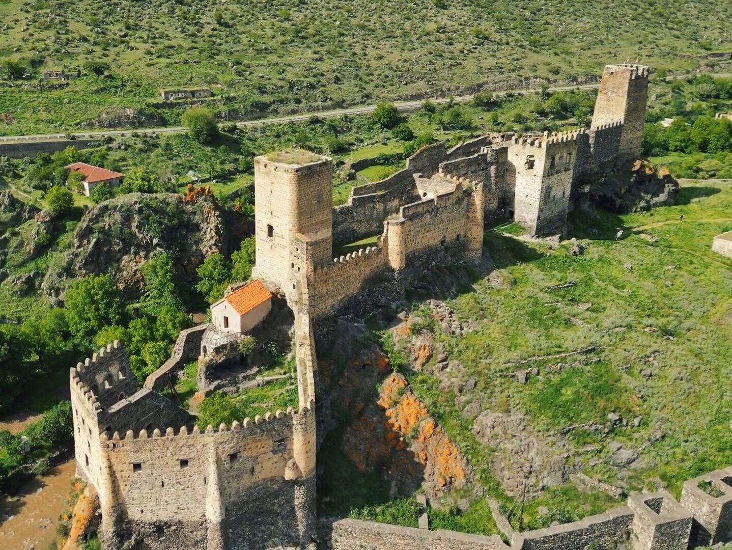 Un castillo medieval con múltiples torres y una muralla fortificada, rodeado de colinas verdes y un pequeño edificio con techo rojo.