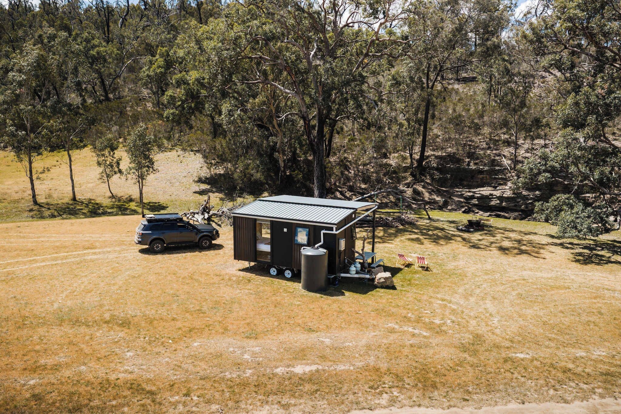 A small cabin with a metal roof, a vehicle parked outside, and a grassy area with trees in the background.