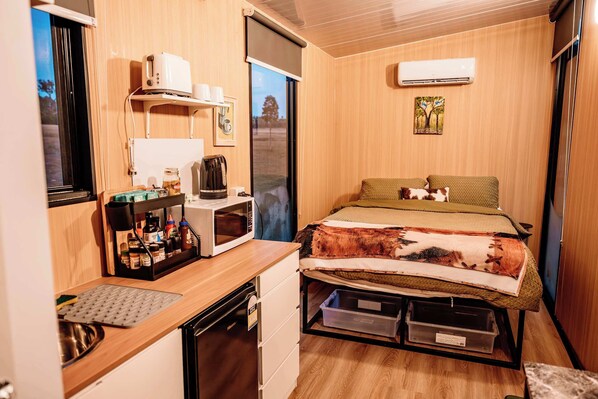 A compact room with a kitchenette, a bed with a blanket, a window, and an air conditioning unit. - Hermano Farm Stay by Tiny Away (Turill)