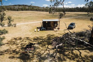 A rustic cabin with a metal roof, surrounded by a dry grassy field, a fire pit, and a parked vehicle. - Hermano Farm Stay by Tiny Away (Turill)