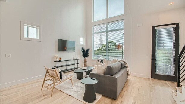 Uma sala de estar moderna com um sofá cinzento, uma mesa redonda pequena, uma cadeira de vime e uma janela ampla com persianas.