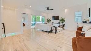 A spacious living room with a white sofa, a brown armchair, and a coffee table. There is a ceiling fan, a TV mounted on the wall, and a large window providing natural light. - 24- Home w/  Rooftop- The Music City Collection (Berry Hill)