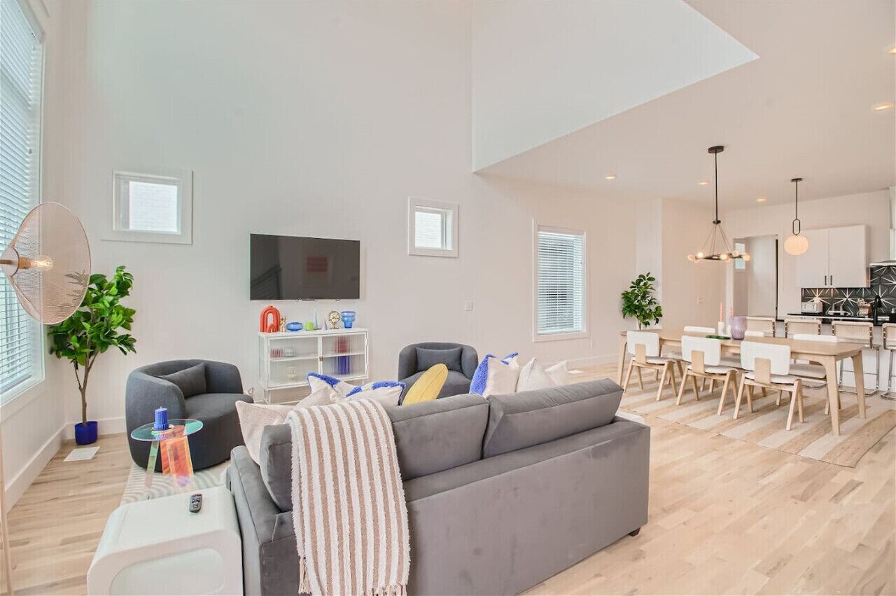 A spacious living area with a gray sofa, a small round table, and a TV. There is a dining area with a wooden table and chairs.