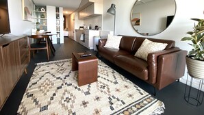 A modern living room with a brown leather sofa, a coffee table, and a patterned rug. There is a kitchen in the background with white cabinets and a dining area with a table and chairs. - Sophisticated Lakeview Apt + Parking (Canberra)