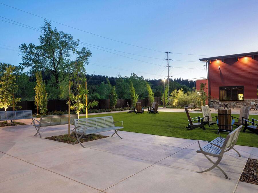 An outdoor patio area with benches, a fire pit, and a red building in the background.