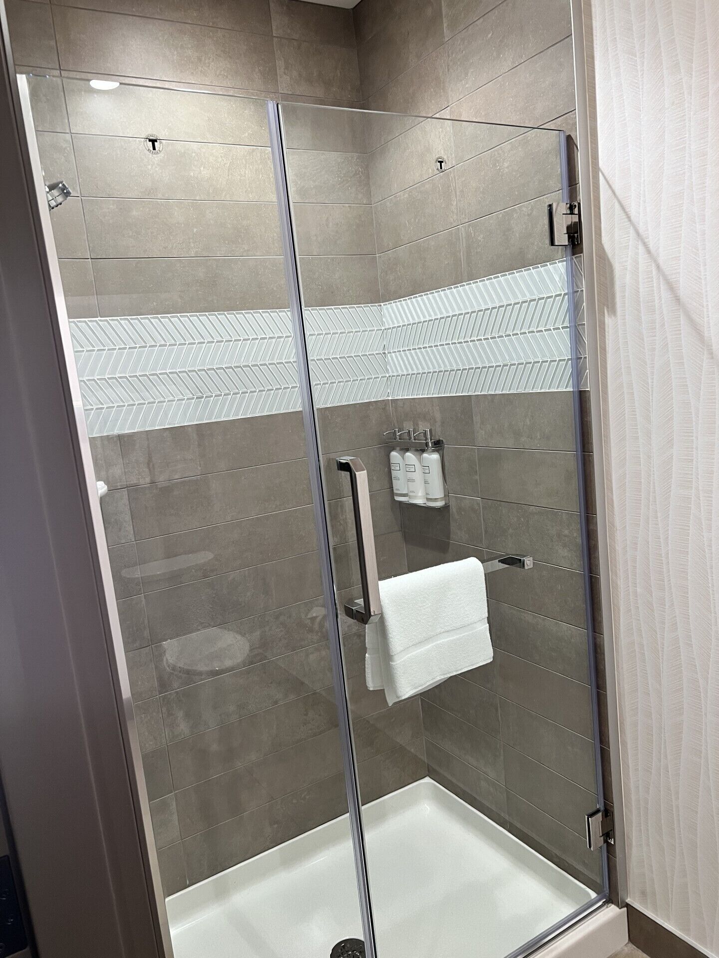 Una ducha moderna con puerta de vidrio, azulejos grises y una toalla blanca colgada en un perchero.