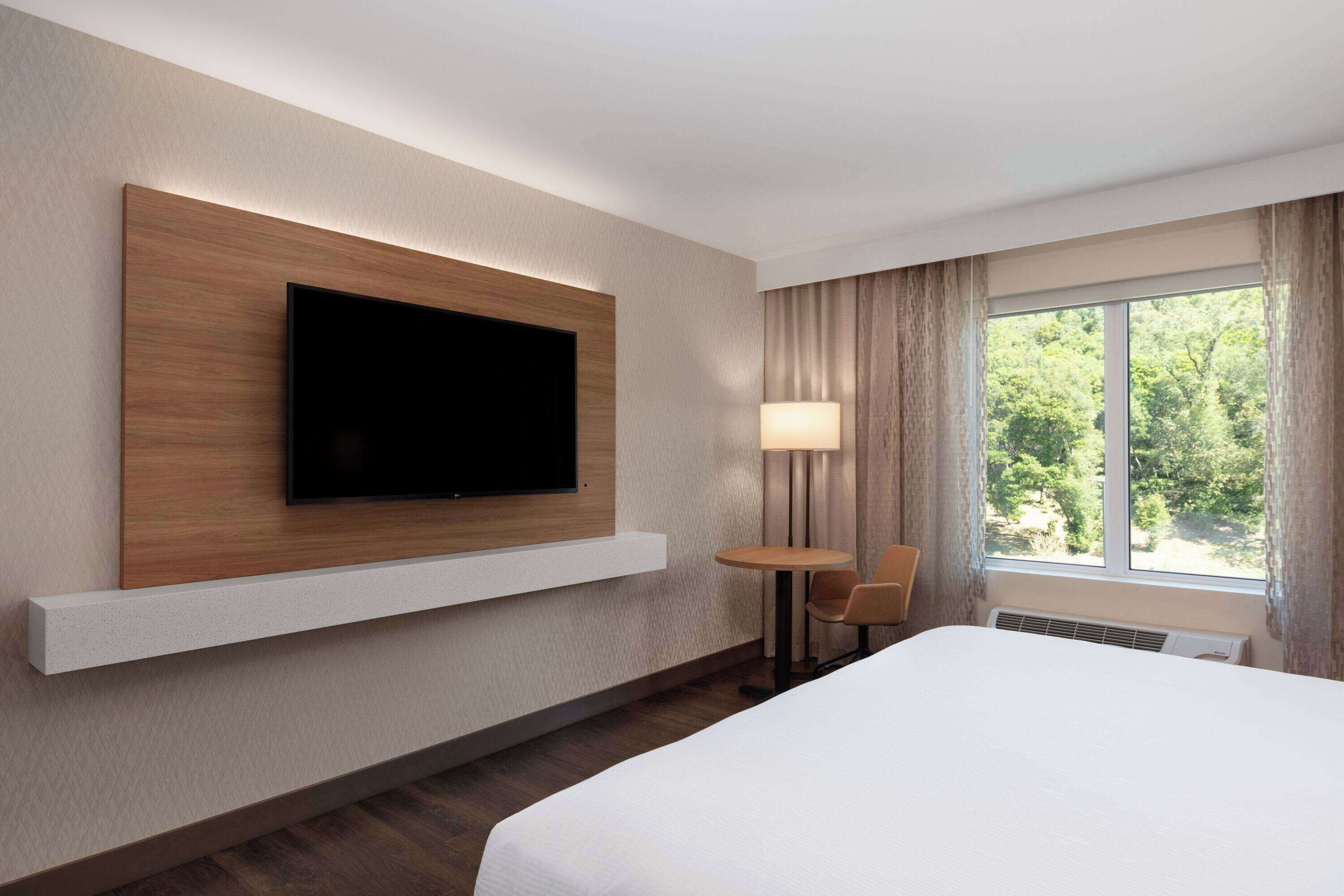 A hotel room with a large bed, a flat-screen TV mounted on a wooden panel, a small table with a chair, a window with curtains, and a lamp.