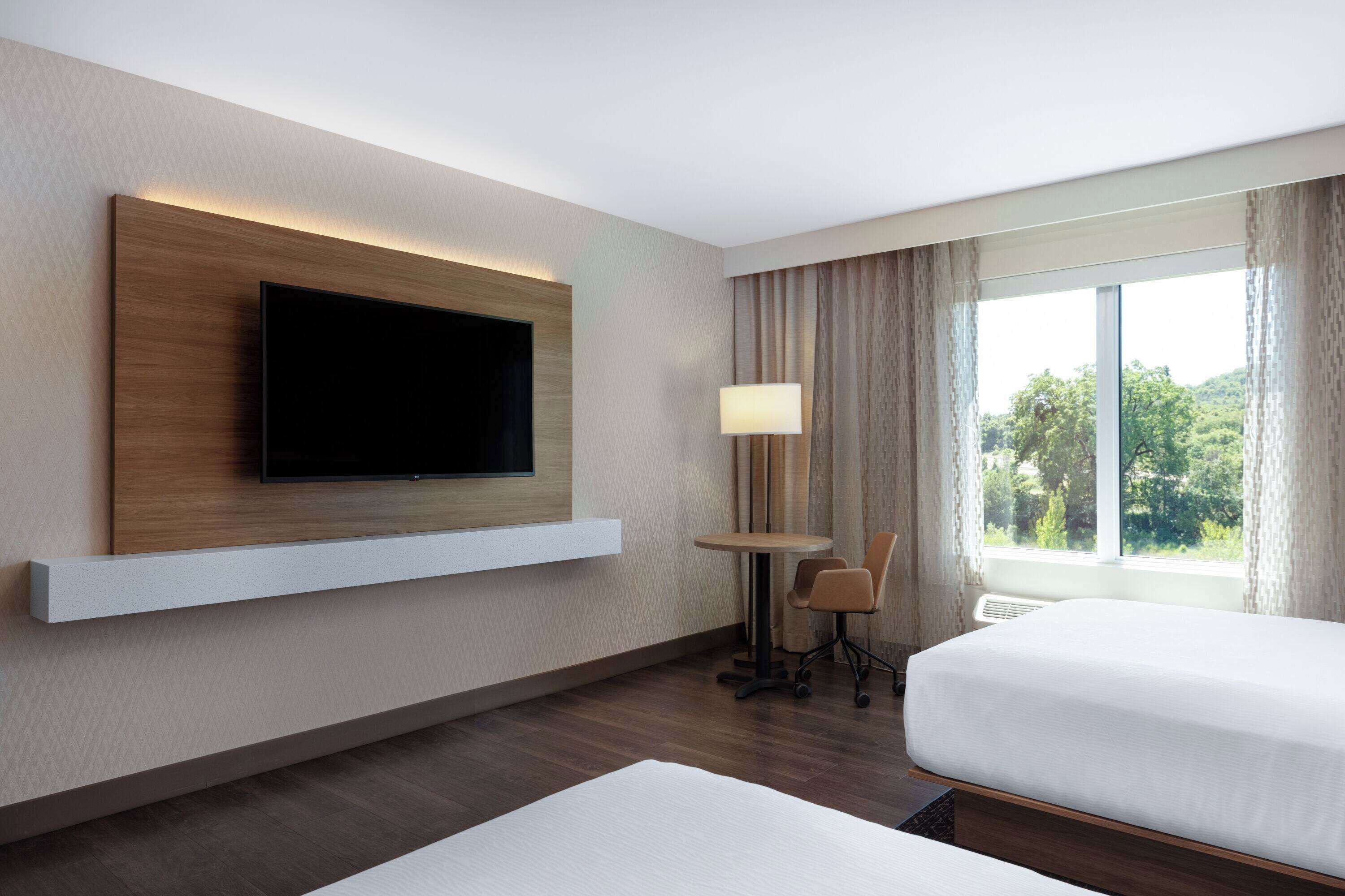 A hotel room with a large flat-screen TV mounted on the wall, a small round table with two chairs, a bed with white bedding, and a window with curtains.