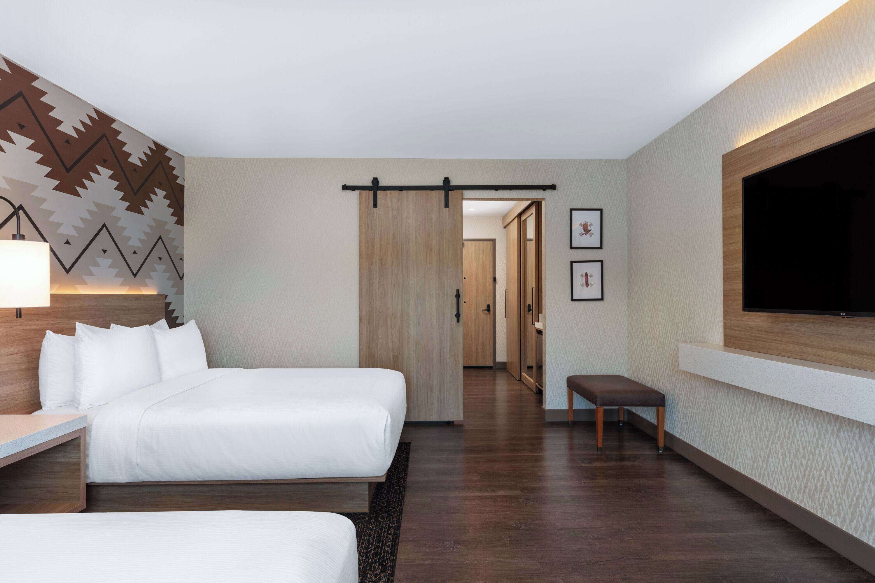 Habitación de hotel con una cama grande, un escritorio, un banco, un televisor montado en la pared y un papel tapiz decorativo.