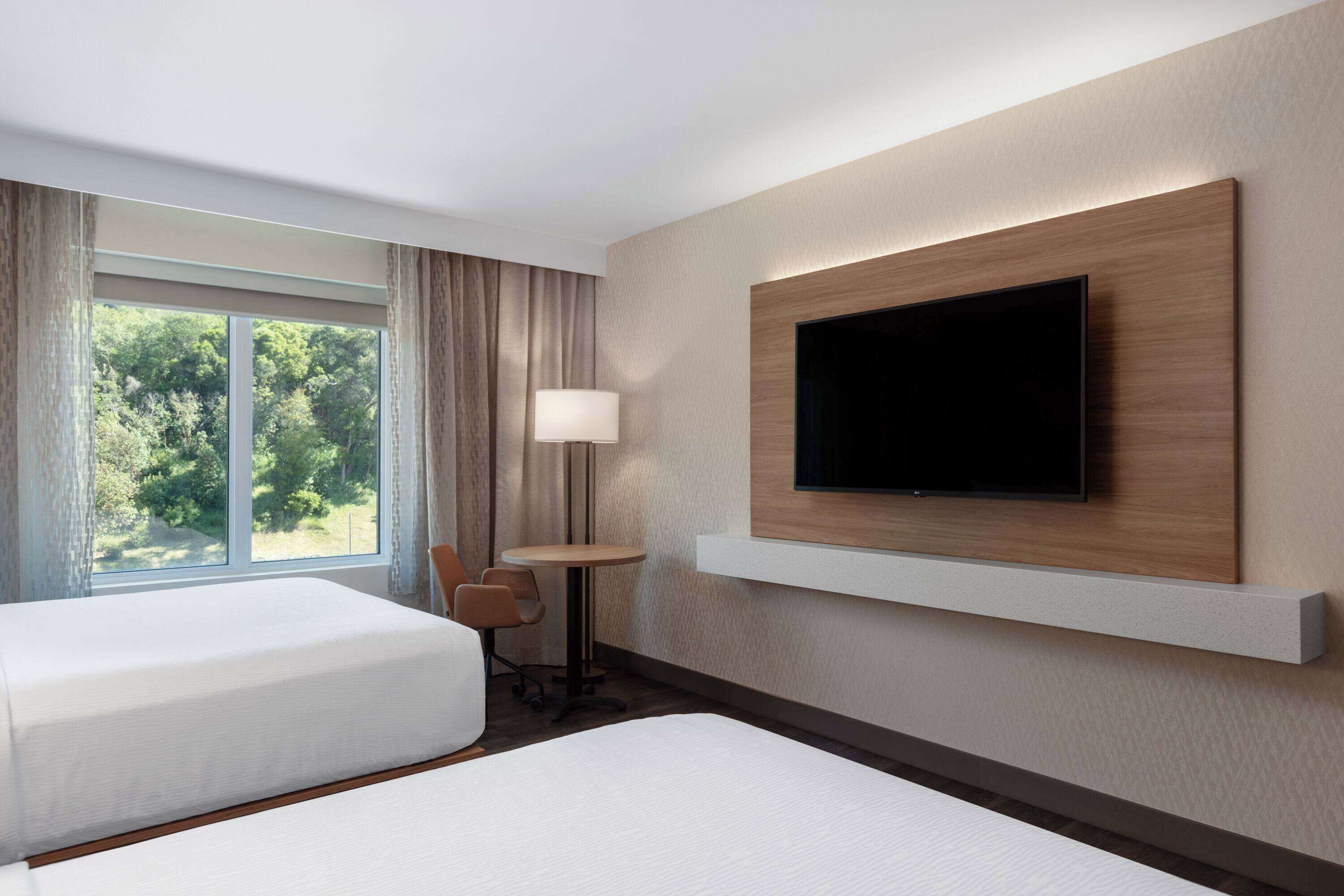 A hotel room with two beds, a TV mounted on the wall, a small table with a lamp, and a view of greenery through the window.