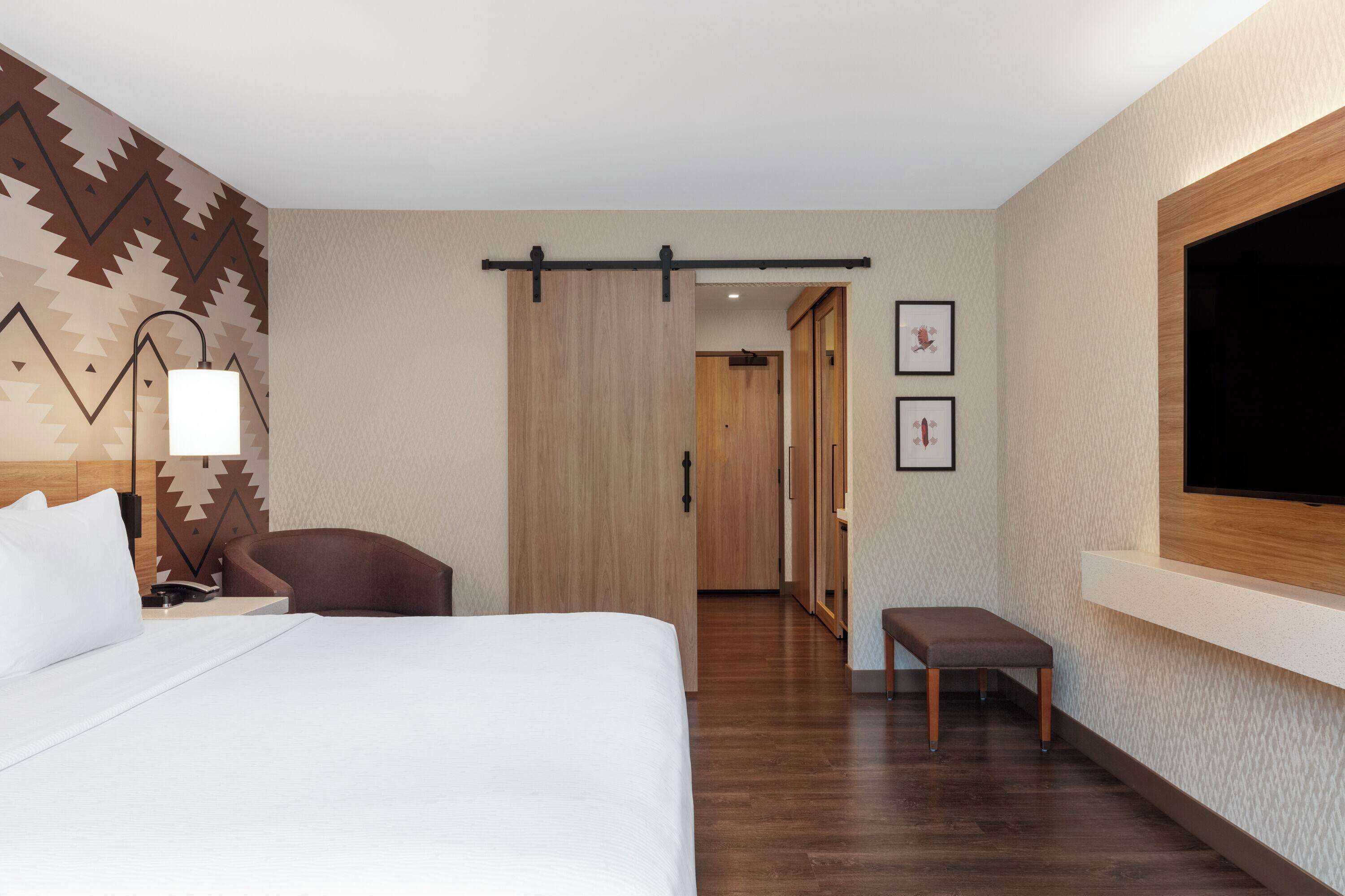 Habitación de hotel con cama, escritorio, silla, banco, televisión y puerta corredera.