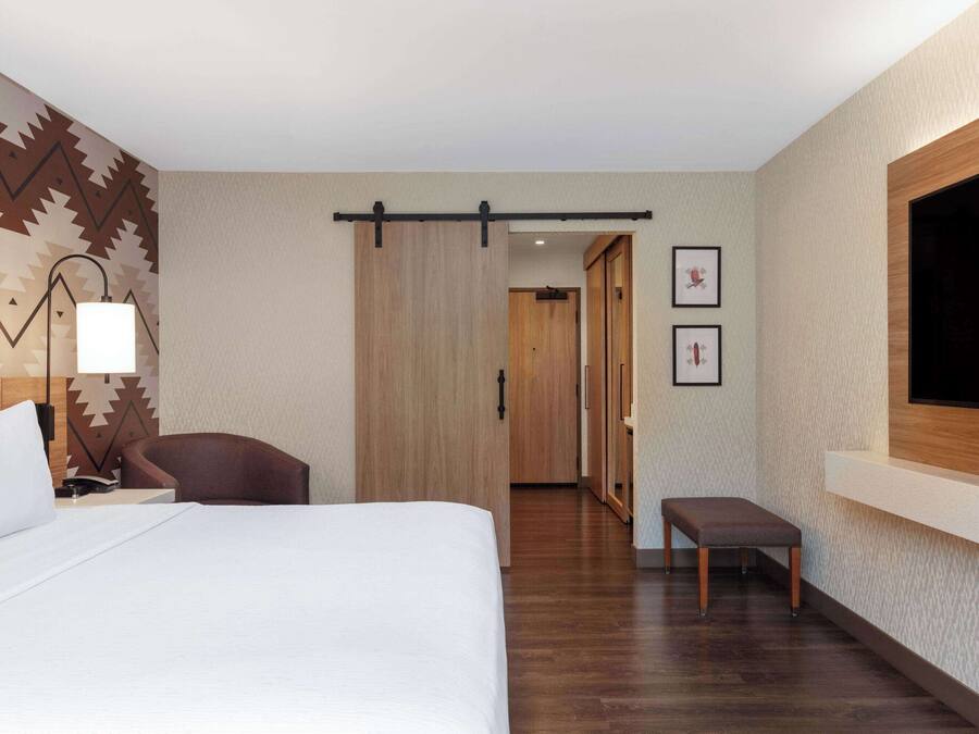 A hotel room with a bed, a desk, a chair, a bench, a TV, and a sliding door.